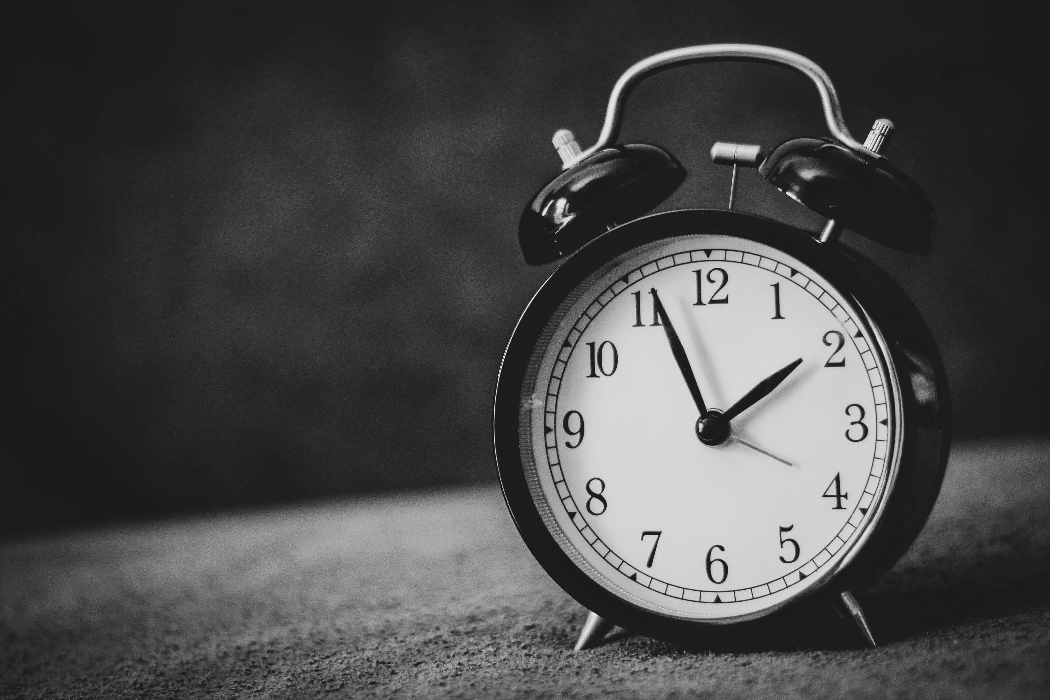 A black-and-white photograph of a vintage-style analog alarm clock showing approximately 7:00, set against a dark, textured background.